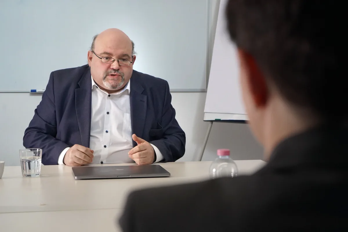 Ein älterer Mann mit Brille sitzt hinter einem Tisch, vor sich hat er einen Laptop.